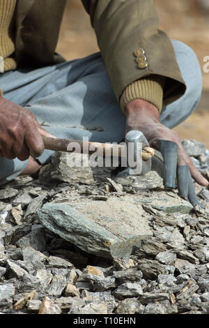 Man, stone breaking, using a handheld hammer or mallet head to chip smaller stones, all of near identical size towards local road construction and mixing to make concrete. Note, 'homemade', finger protection! Northern India. Stock Photo