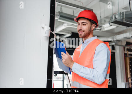 cheerful fireman in helmet checking fire alarm while holding clipboard Stock Photo