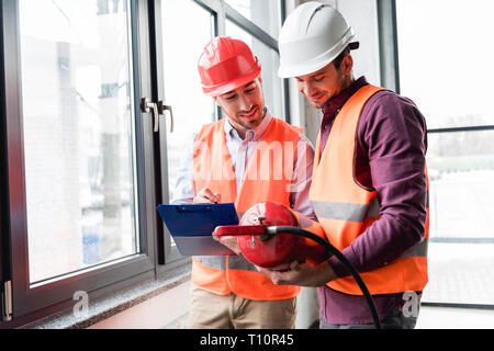 cheerful firemen in helmets standing and looking at red extinguisher Stock Photo