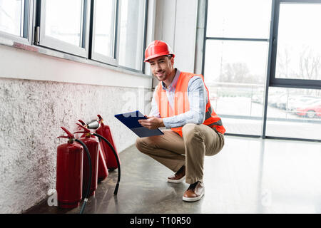 happy fireman in helmet holding clipboard and pen while looking at camera near extinguishers Stock Photo