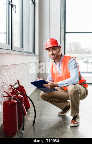 cheerful fireman in helmet holding clipboard and pen while looking at camera near extinguishers Stock Photo