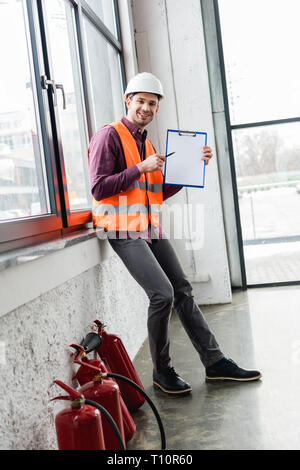 cheerful fireman in helmet and uniform holding clipboard with blank paper near extinguishers Stock Photo