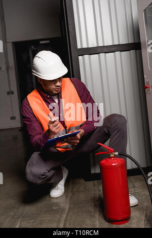 thoughtful firefighter in helmet sitting and looking at extinguisher near fire panel Stock Photo