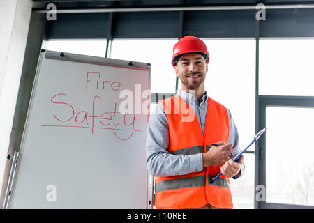 cheerful firefighter in helmet holding clipboard and pen while standing near white board with fire safety lettering Stock Photo