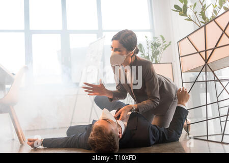 terrified businesswoman in mask sitting near colleague lying on floor in office with smoke Stock Photo