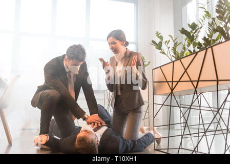 terrified businesswoman screaming near colleague lying on floor while man giving first aid  in office with smoke Stock Photo