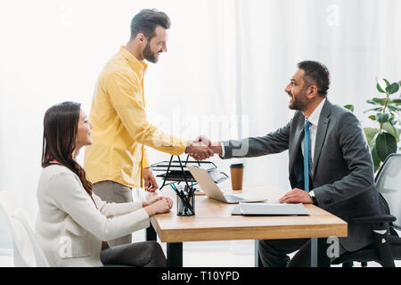advisor and investor shaking hands over table wile woman sitting in office Stock Photo