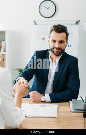 handsome advisor in suit shaking hands with investor at workplace Stock Photo