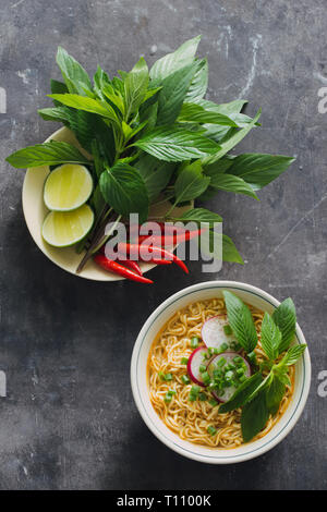 Instant noodles in bowl with fresh herbs, garnish of cilantro and Asian basil, lemon, lime on dark stone background Stock Photo