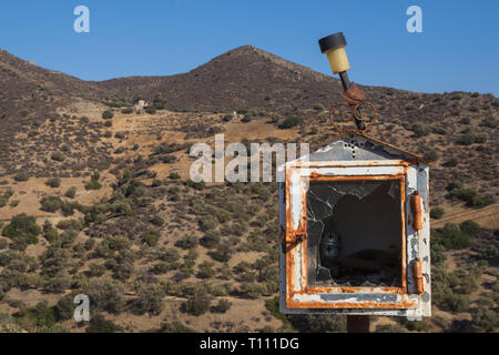 Shrine with a broken glass and a candle inside. Solar lamp on the roof. Mountains with olive trees in the background. Blue sky. Central Crete, Greece. Stock Photo