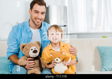father and preschooler son sitting on couch and holding teddy bears at home Stock Photo