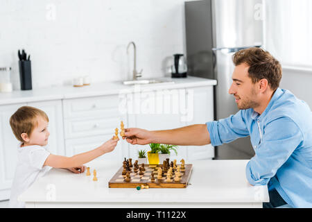 father and preschooler son holding chess pieces while sitting at table and playing chess at home Stock Photo