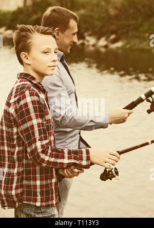 Teenage boy and father fishing together on freshwater lake from shore Stock Photo