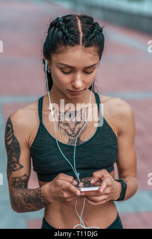 Close-up, a girl in hands of smartphone, tanned skin, cat tattoo, application select playlist of music for training, in summer in city. Active Stock Photo