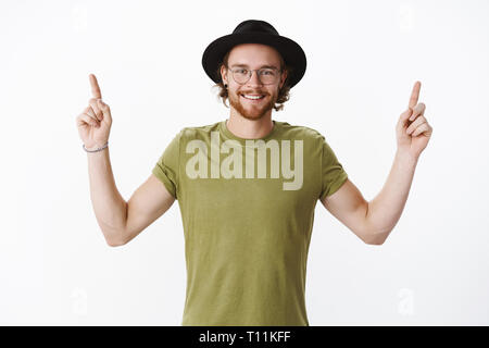 Waist-up shot of good-looking happy charismatic redhead bearded guy in hat and olive t-shirt raising hands pointing up joyfully as recommending good Stock Photo