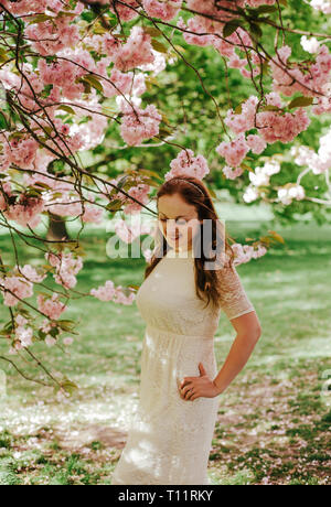 beautiful cute brunette girl bride in cream lace dress, with decoration on the hair, near the tree blossoms with pink flowers, garden, greenwich park Stock Photo