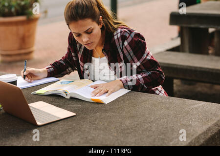 Female student sitting at college campus and making notes. Woman student studying at university campus with a laptop in front. Stock Photo