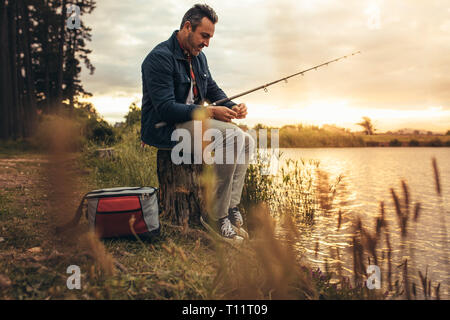 Man sitting on a tree trunk beside a lake with his fishing rod. Man enjoying his leisure time fishing in a lake. Stock Photo