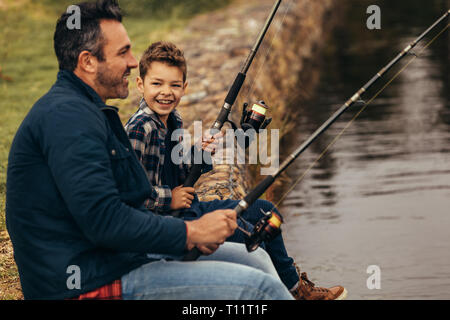 Side view of a man sitting on the banks of a lake and fishing with his kid. Close up of a man winding the reel of his fishing rod while his son looks  Stock Photo