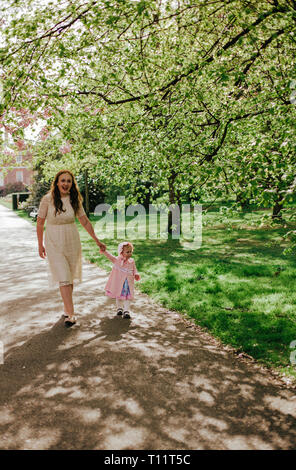 Beautiful woman in cream lace dress with daughter in pink dress walking in park outdoors green tree, spring. Family on nature in Greenwich park, UK Stock Photo