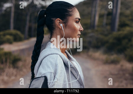 Side view of young woman with earphones standing outdoors after morning run. Female taking a rest after outdoor workout. Stock Photo