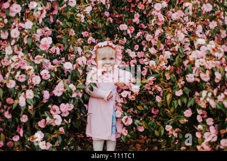 Pretty little girl wearing pink bonnet and pink dress in blossom bush of pink camelia in sunny spring day Stock Photo