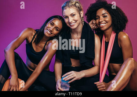 Three young women resting after exercising in fitness studio.  Diverse group of female in sports clothing sitting on colored background looking at cam Stock Photo