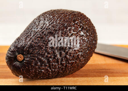 Whole ripe dark brown avocado bilse variety on wooden cutting board, blurred knife in background Stock Photo