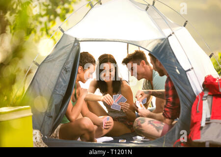 Group of young people on camping trip in countryside sitting in tent and playing cards Stock Photo