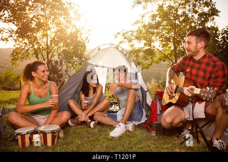 Positive young people on camping trip sitting next tent and playing guitar Stock Photo