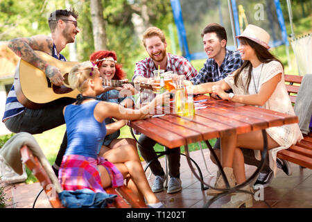 Smiling happy people tapping with glasses of beer while tattooed guy plays guitar outdoor in summer Stock Photo