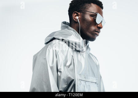 Handsome african man in silver hooded shirt, sunglasses and earphones against white background. Funky young african american man. Stock Photo