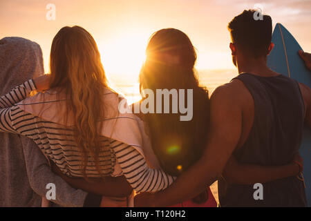 Rear view of a group of friends standing together on beach holding each other. Man standing with his women friends at the beach looking at the sunset Stock Photo