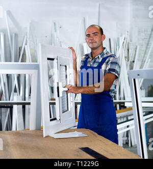 Glad workman in blue overalls demonstrating pvc window in workshop Stock Photo