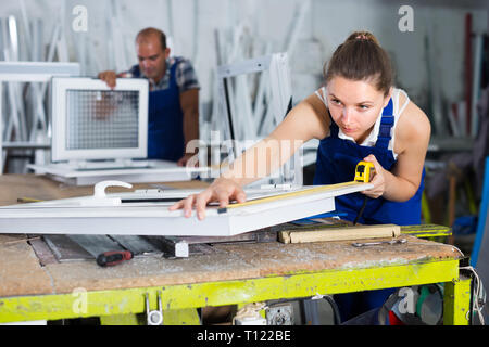 Woman 20-30 years old is checking dimensions of finished plastic window using measuring tape at factory. Stock Photo