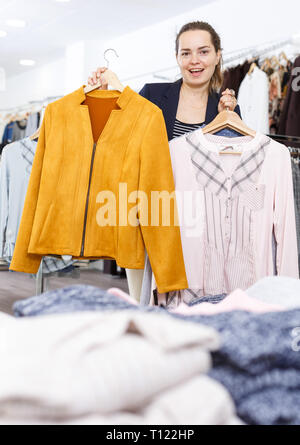Young cheerful woman demonstrating clothes on hangers in clothing boutique Stock Photo