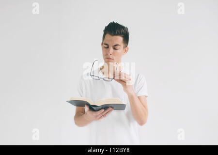 nerd with glasses and a white t-shirt is reading a book on a white background. A man likes to read books Stock Photo