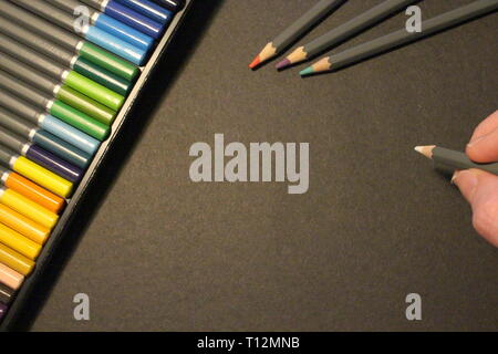 A photograph of a neat set of coloring pencils, three out ready on black paper, with a hand holding a white coloring pencil and copy space to add text Stock Photo