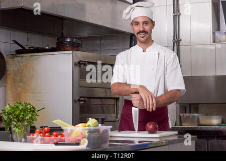 one young man, dressed in chef uniform while holding a knife on a cutting board. Posing, looking to camera. Indoors, in a professional restaurant kitc Stock Photo