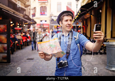 Young tourist man on streets of old town, having map, vintage photo camera and taking urban break selfie with his smart phone camera Stock Photo