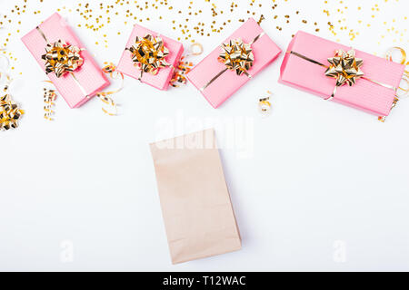 Plain brown paper bag next to festive pink gift boxes with golden bows, ribbons and glitters on white background, view from above. Stock Photo