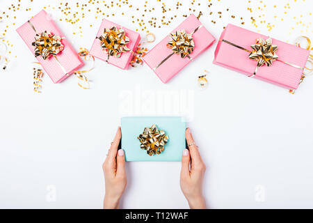 Festive flat lay composition woman's hands giving blue gift box among pink presents with golden bows and ribbons on white table, view from above. Stock Photo
