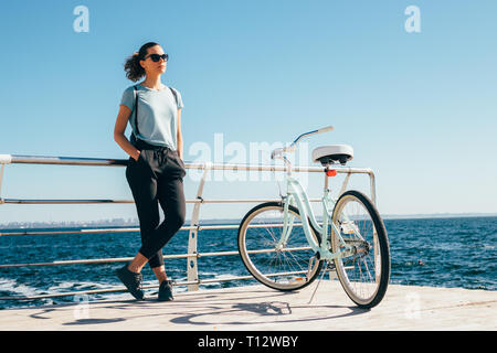 Young woman standing at the background of blue sea near her shiny cruiser bicycle. Female wearing backpack and casual outfit with hands in pockets res Stock Photo