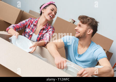Young husband and wife moving to new place sitting near box packing things in bubble wrap talking laughing cheerful Stock Photo