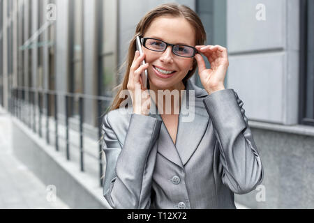 Young businesswoman wearing eyeglasses standing on the city street near building calling to friend on smartphone looking camera smiling playful close- Stock Photo