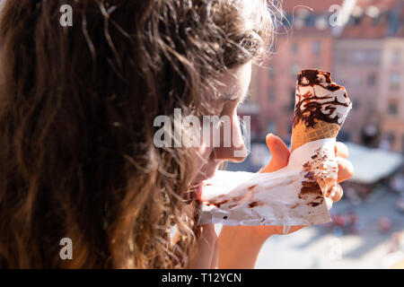 Woman eating chocolate ice cream gelato cone paper licking with background of Warsaw, Poland old market square historic buildings in city during sunny Stock Photo