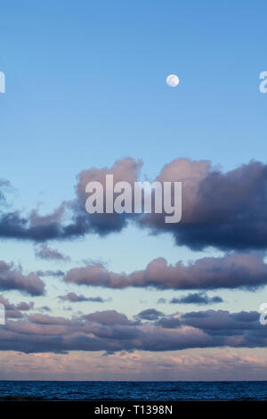 Impressive sky at sunset with a full moon above and layers of clouds tinged with pink that slope down to the horizon over the dark blue sea Stock Photo