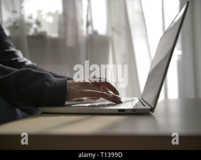 female hands are typing on the keyboard of the laptop on the background of the window Stock Photo