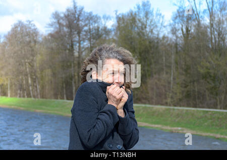 Woman snuggling into her jacket on a cold day as she enjoys a walk in the fresh air alongside a lake or river Stock Photo