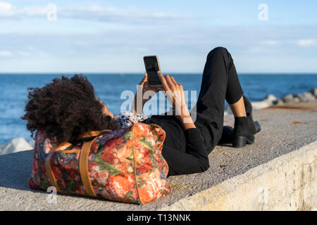 Woman dressed in black relaxing on a wall in front of the sea using smartphone Stock Photo